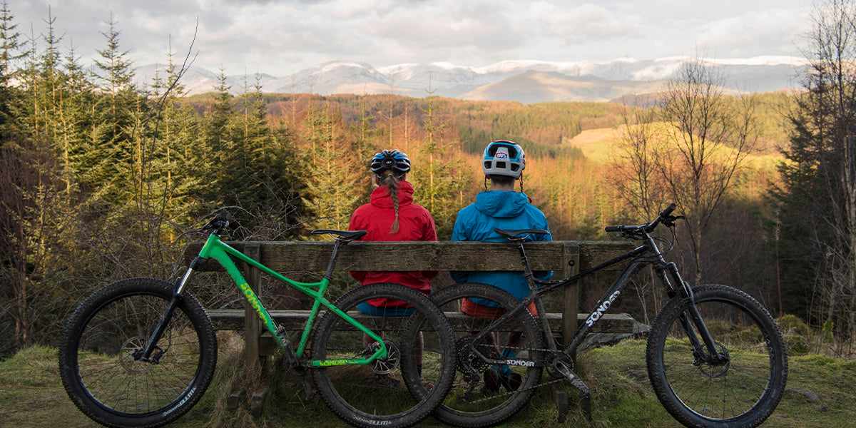 Two mountain bikers sitting on a bench in front of their bikes and looking out at the forest and mountains