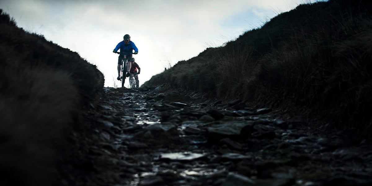 Two people riding fast down a rocky descent on mountain bikes during a wet day
