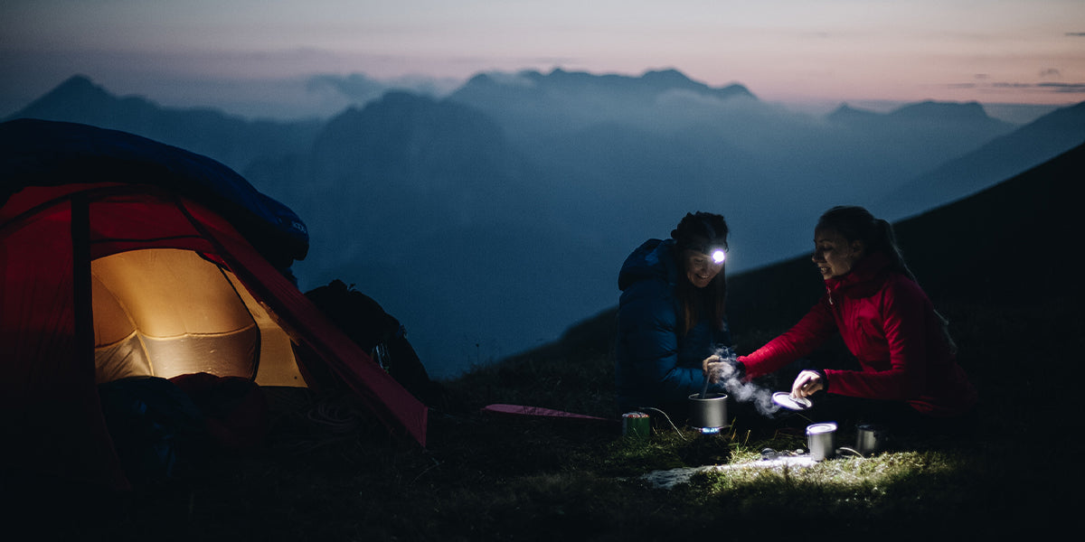 Two women cooking dinner next to their tent at dusk in the Slovenian mountains
