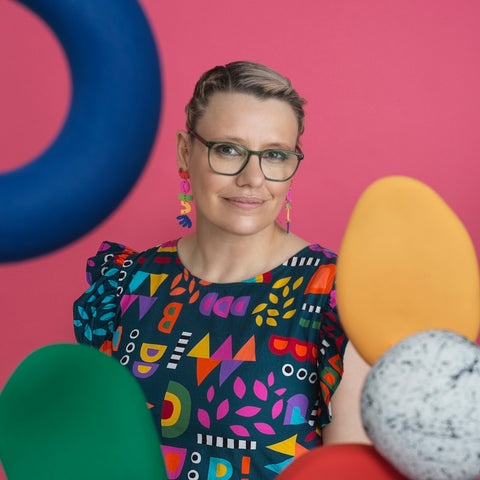 Photo of a woman sitting down with just her head and top half of her body visible. She is wearing glasses and long earrings designed by her. She is also wearing a dress in a bold and bright pattern which is also designed by her. The photograph contains different shapes, which are made from polymer clay in bright colours and are used as props in the photograph.