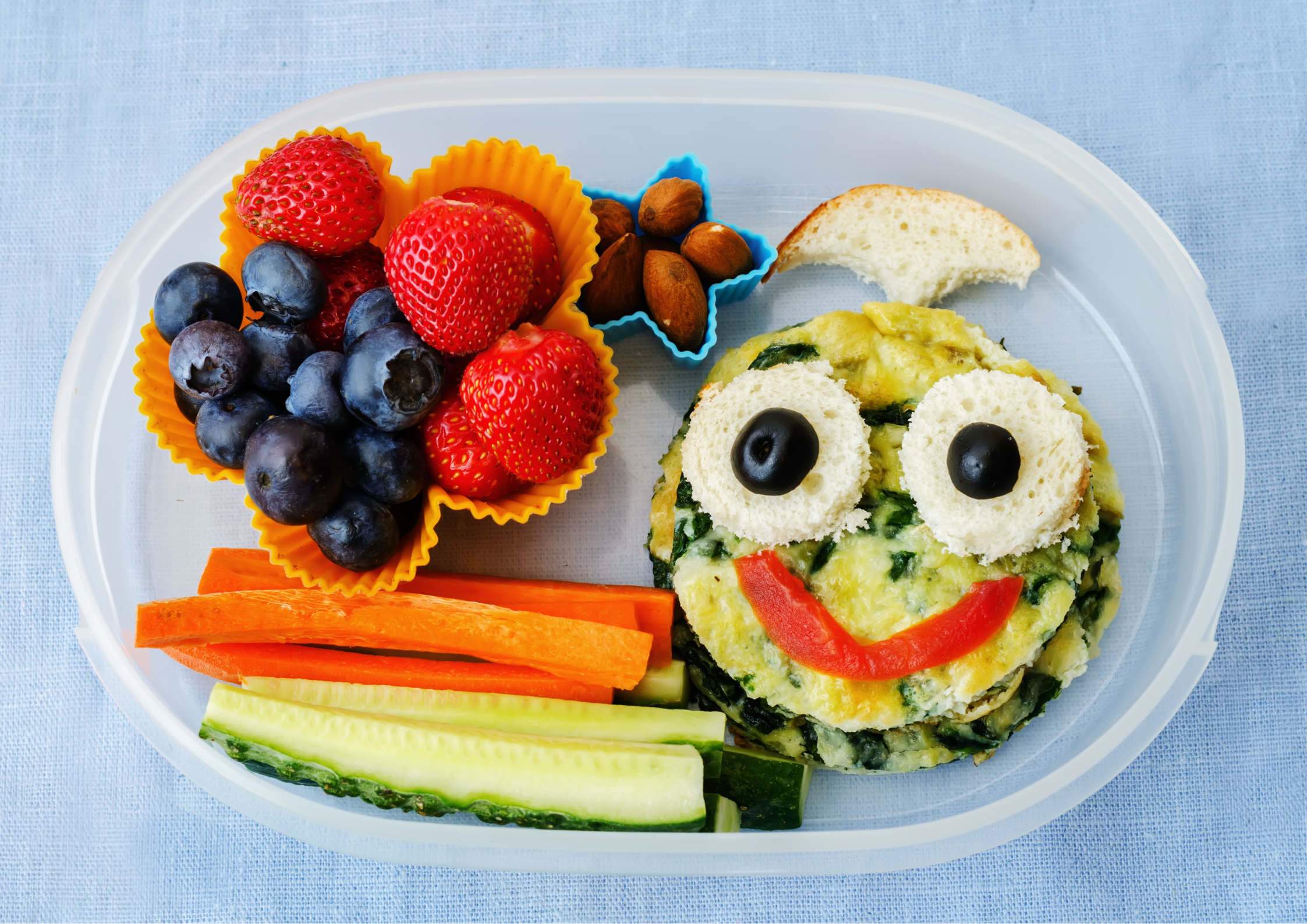 A clear Tupperware with a round omelette decorated to look like a smiling face with cut up vegetables, strawberries and blueberries. 