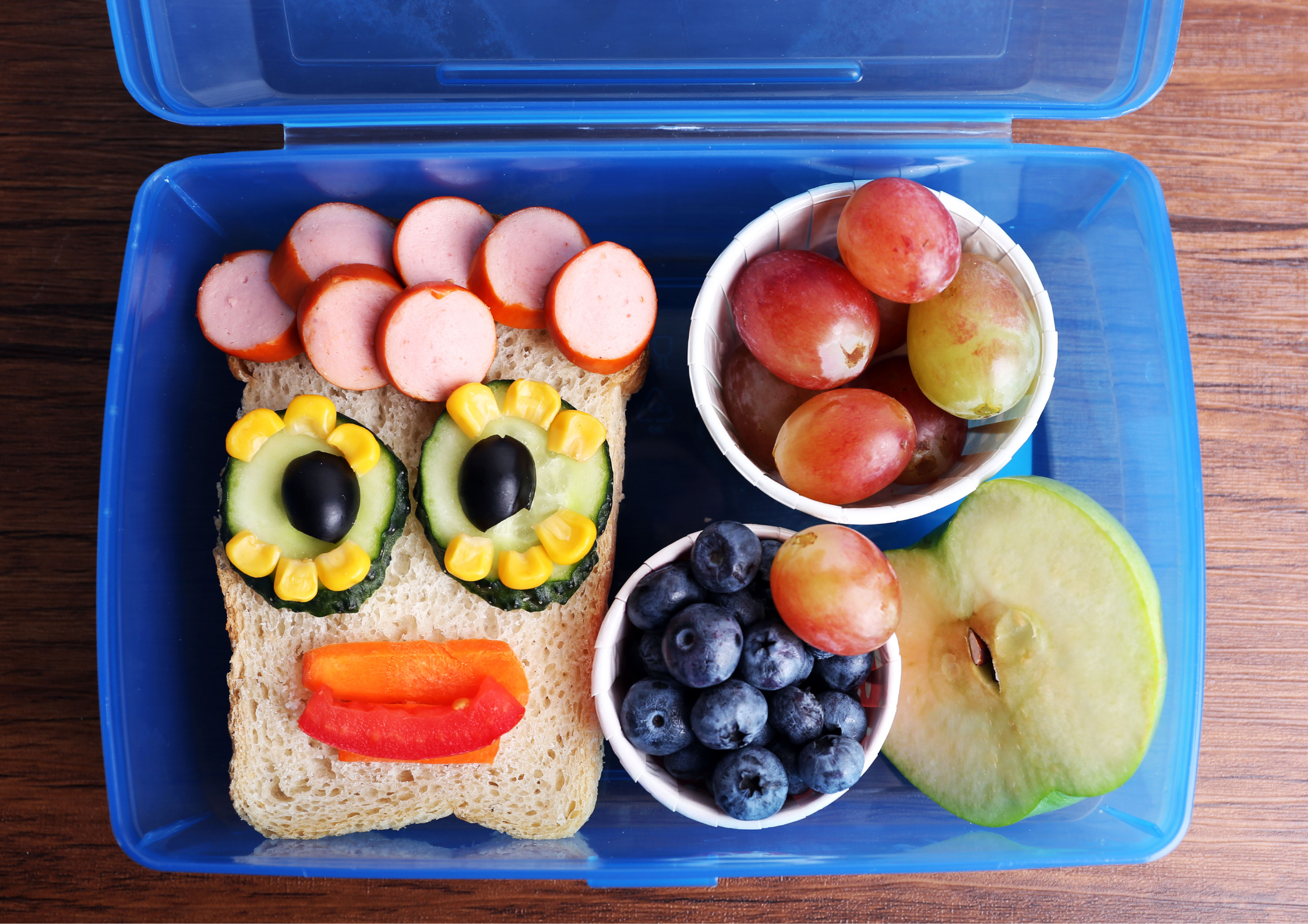 A blue lunchbox with a sandwich decorated to look like a smiley face next to a cup of blueberries, a cup of grapes and half an apple cut up.