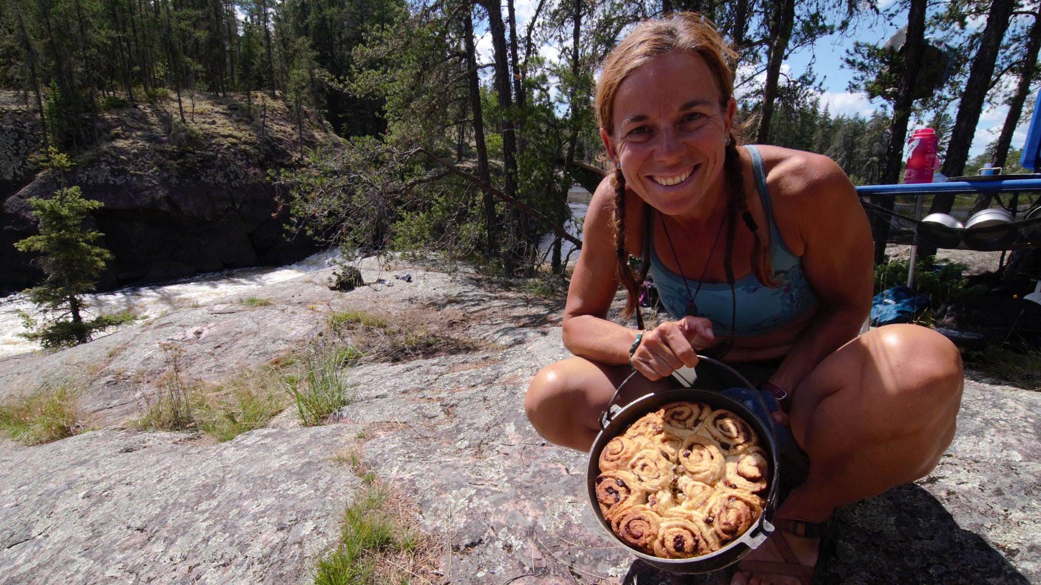 womanoutside crouched on a rock holding out a dutch oven full of cinnamon buns made while camping