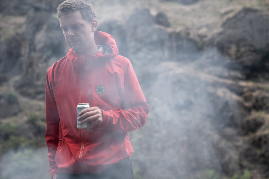 young man in red waterproof jacket by smoke of evening camp fire 