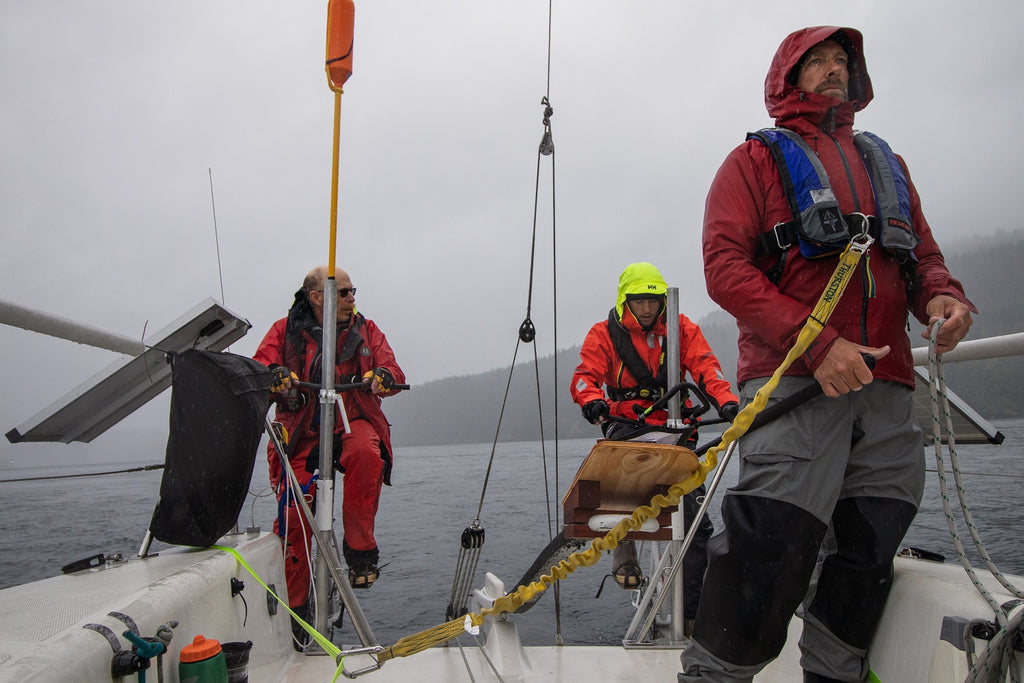 three men in foul weather gear on sail boat. two at back are peddling 