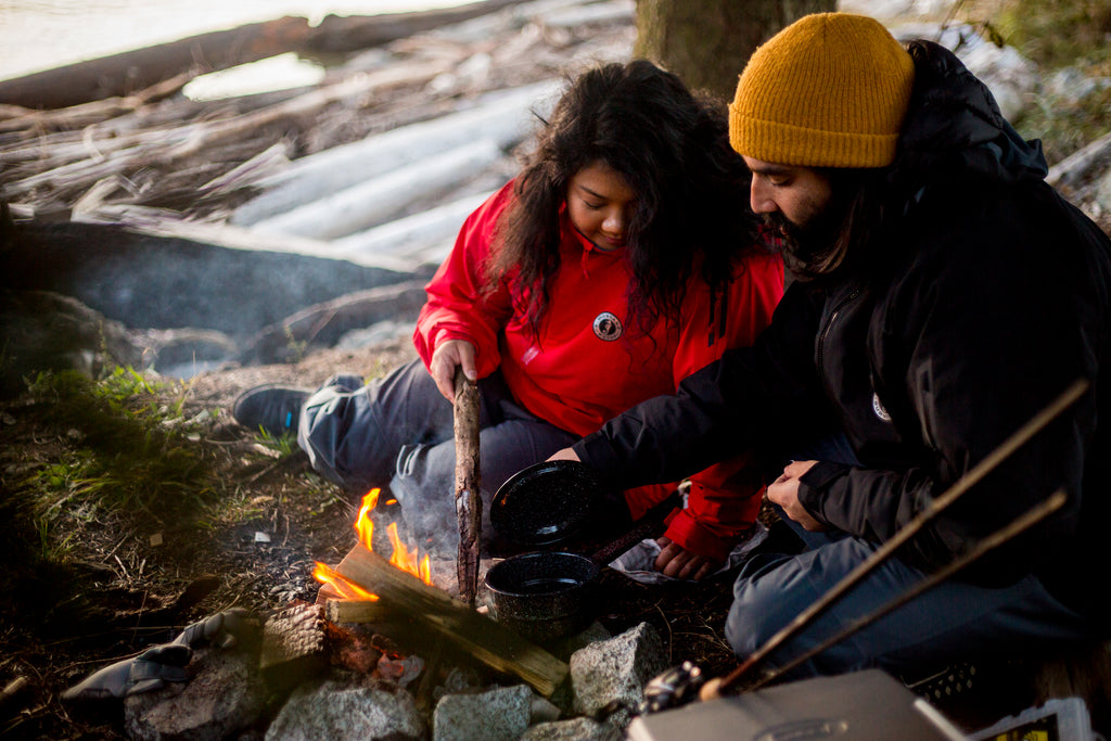 young couple in waterproof jakets cooking over open camp fire by the water