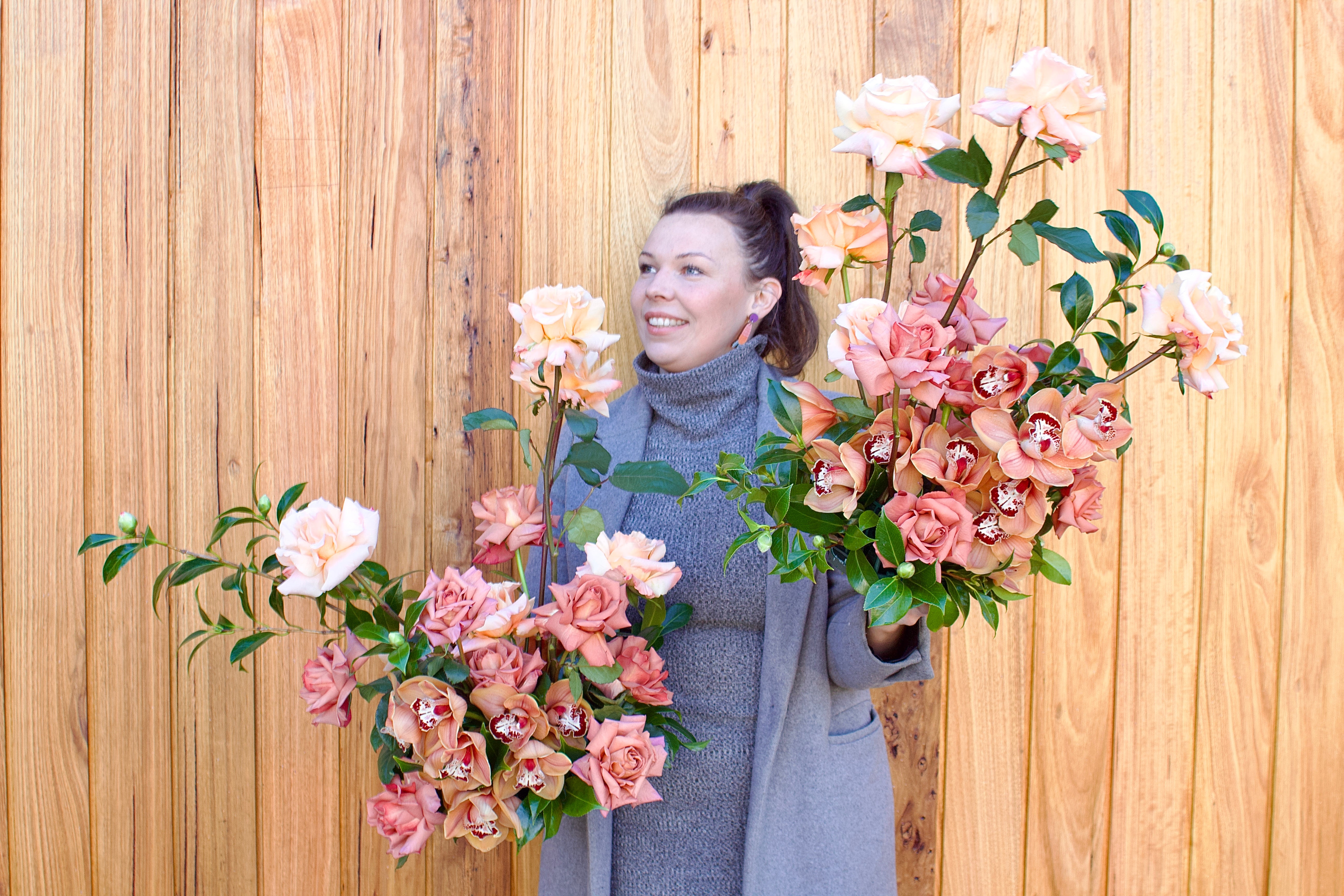 Creative director Emma Schreenan holding two floral arrangements featurimg cymbidium orchids, reflexed roses and camellia.