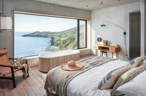 Photograph of relaxed double bedroom with natural linen, poured concrete and floorboards. Complete with freesanding bathtub and stunning view out onto the coast.