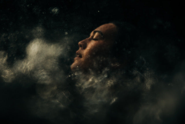 Underwater photograph close up of swimmers face, eyes closed surrounded by blurry air bubbles.