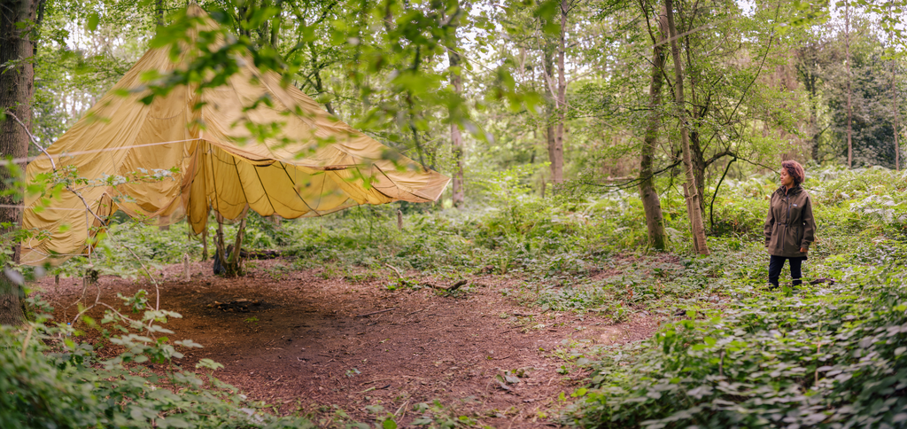 Photo of Beth Collier stood next to a tarpaulin shelter in dense, green woodland (by Danny North)