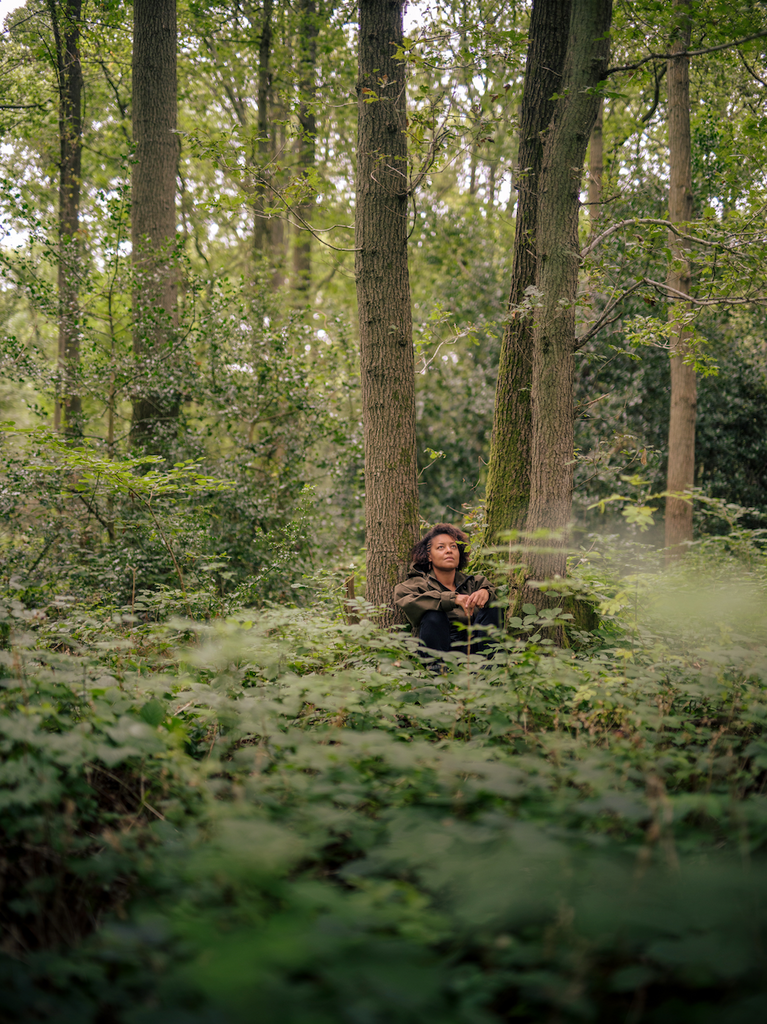 Photo of Beth Collier sitting with her back rested on a tree trunk, surrounded by green woodland (by Danny North)