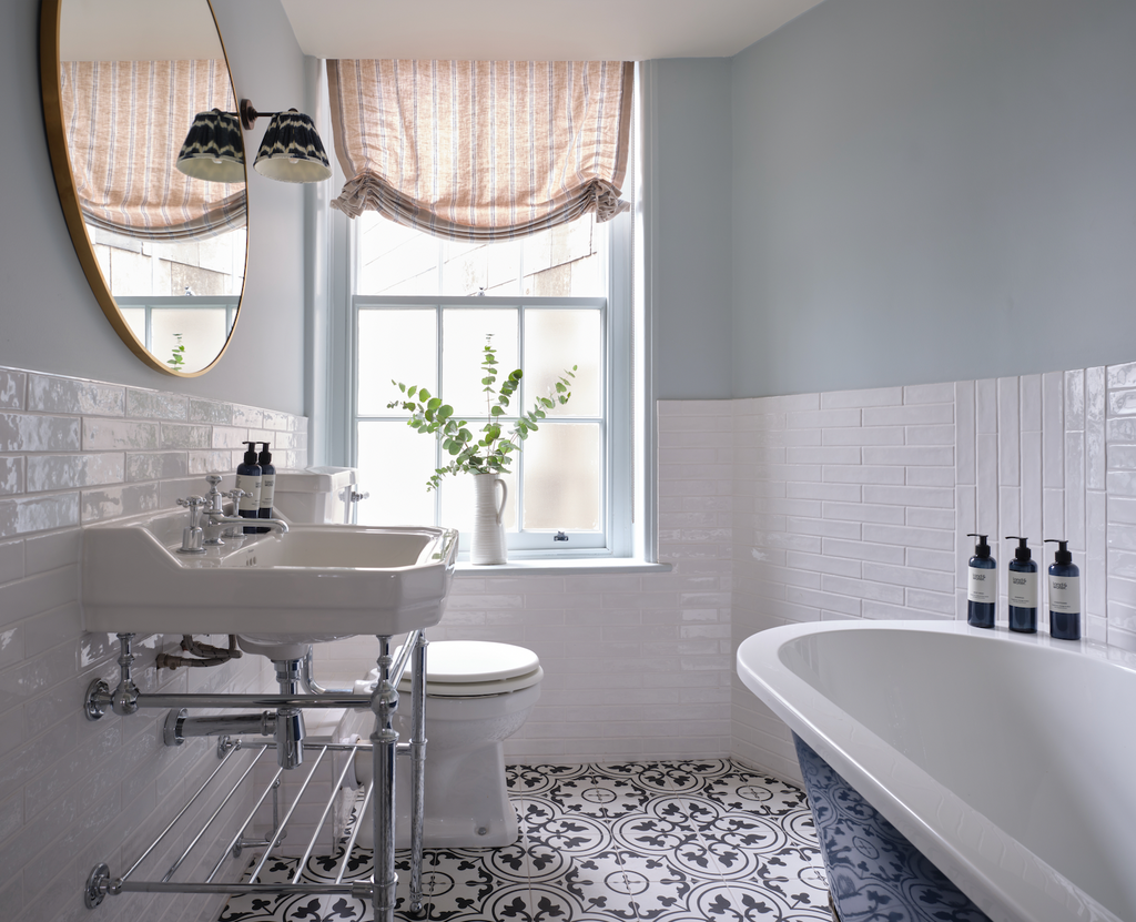 Bright tiled bathroom with sash windown in background, roll-top bath and edwardian bathroom sink in foreground. Stanwell House Hotel.