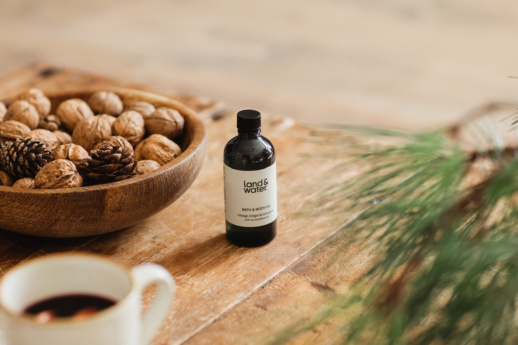 land&water Body Oil product pictured on wooden table with mug of mulled wine and bowl of nuts.