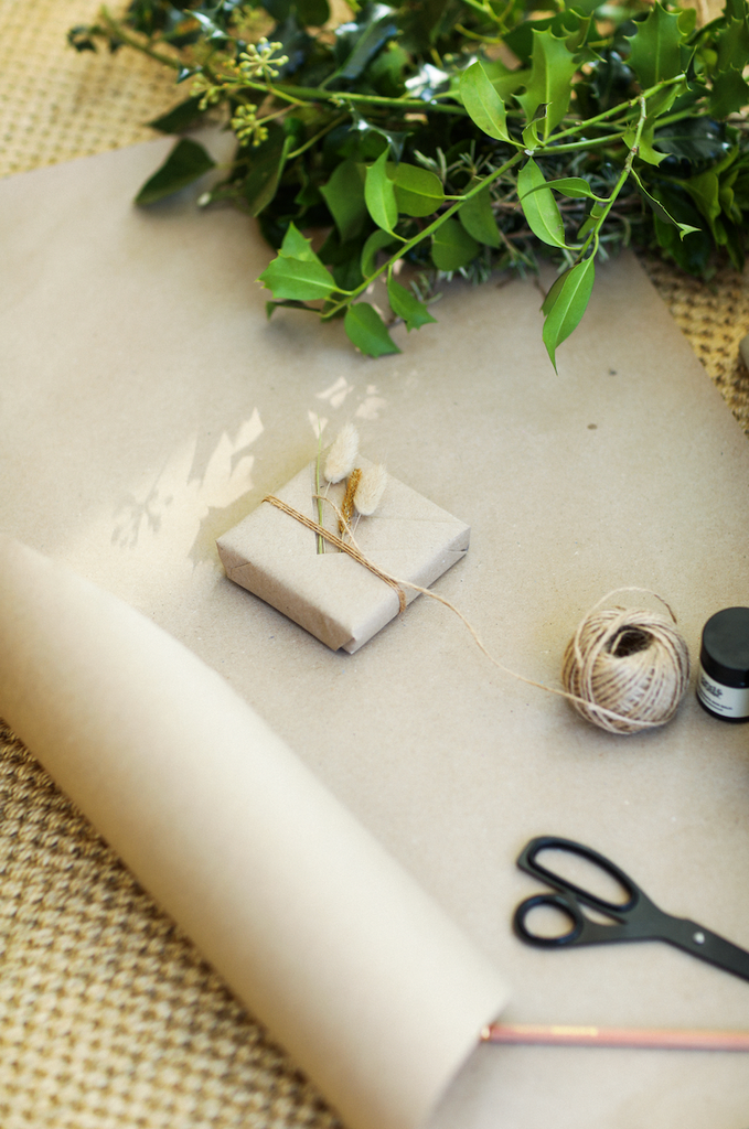 Small present beautifully wrapped in brown paper, string and decorated with natural grasses. Pictured with a roll of brown paper, scissors and holly and ivy sprigs.