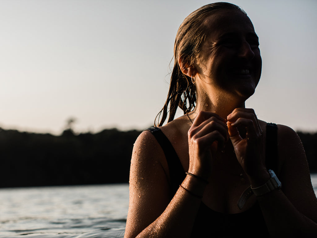 A woman in a swimming costume, rises up from a cool water dip, wet hair and sun shining on her smiling face.