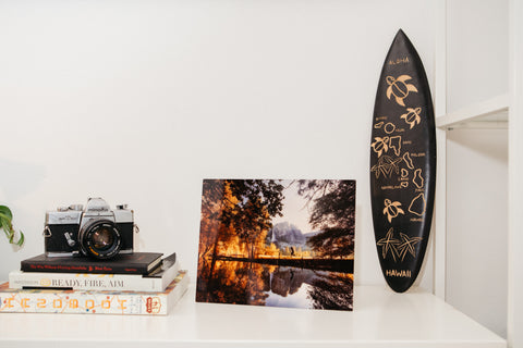 metal print of a fall scene on a lake, hanging next to a surfboard and camera on a desk