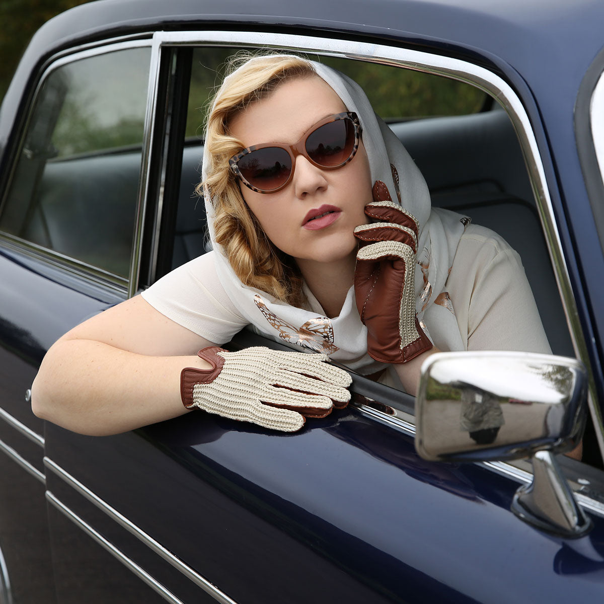 Woman wearing crochet-back leather driving gloves in cognac whilst sitting in a classic car