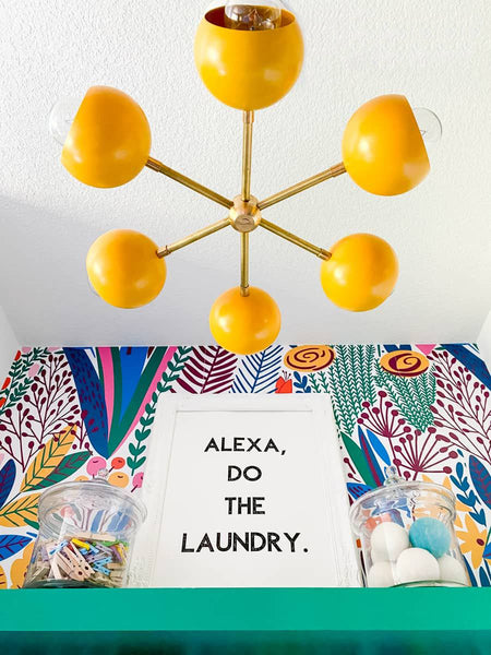 Mustard & Brass Loa Carousel Chandelier in a bold laundry room that features colorful wallpaper, green cabinetry, and humorous laundry-pun accessories.  Lighting  by Sazerac Stitches