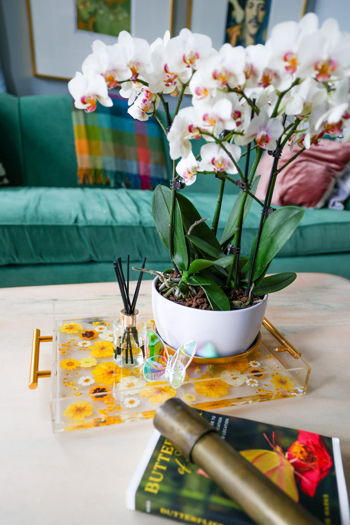 Yellow floral acrylic tray in a living room on a pink marble coffee table with white orchids and trinkets