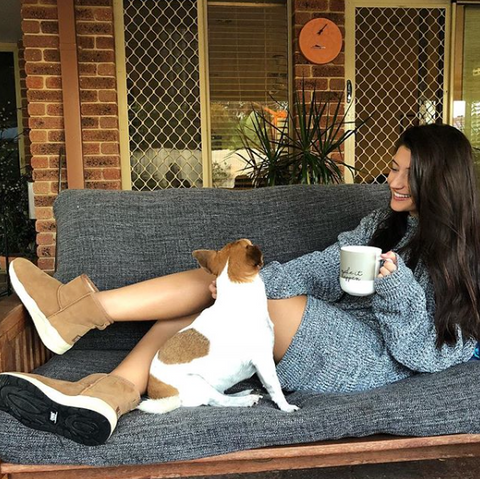 Women wearing her UGG boots with a cup of tea and her pet dog, all on a couch.