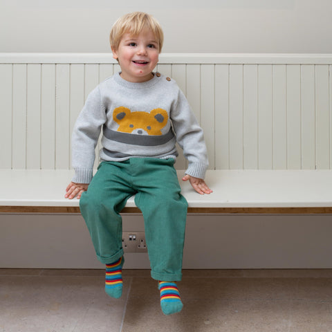 A young boy smiling whilst sitting indoors on a bench wearing Kite's beary knitted jumper.