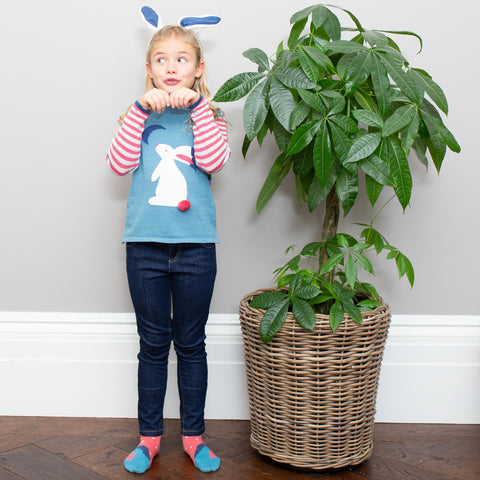 A young girl standing next to a large potted plant, wearing Kite's moongazer jumper and bunny ears. She is posing with her hands under her chin pretending to be a rabbit. 