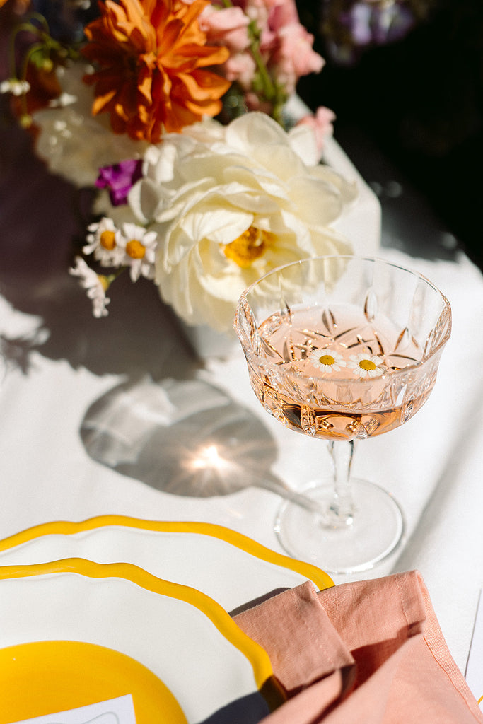 glass of rose with daisy flowers floating in the glass sits on a wedding table in the sun 