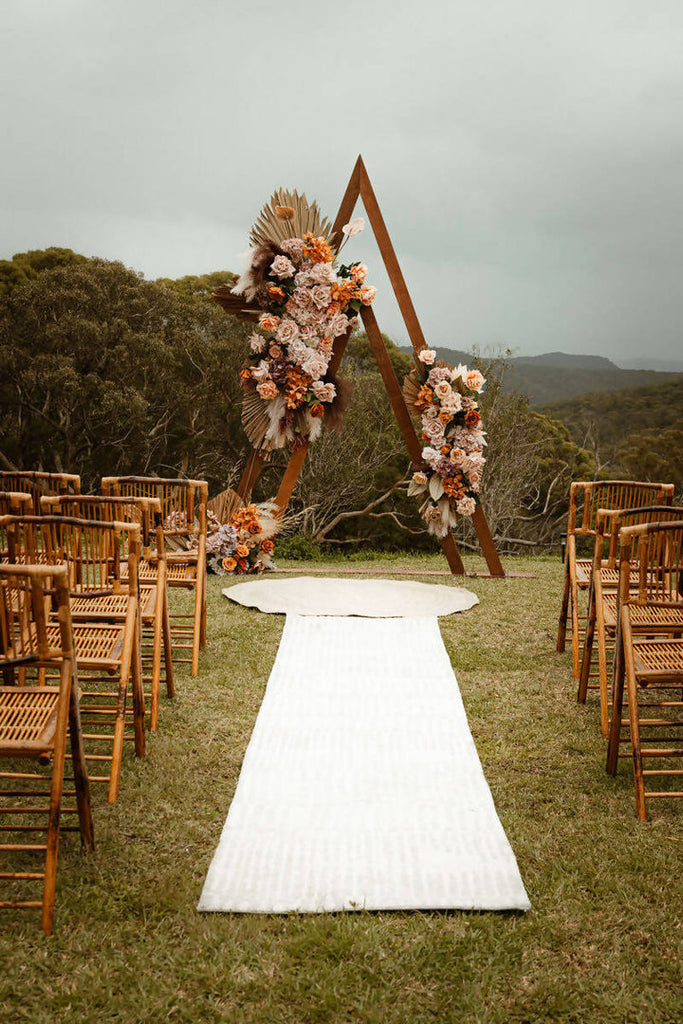 Wooden large triangular arbour decorated with florals stands with rows on wooden chairs creating an aisle with a white rug running down the centre 