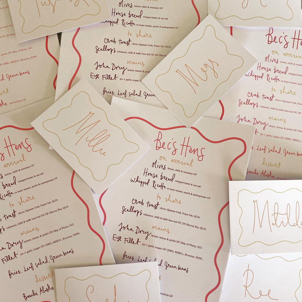 A pile of menus and place cards lay spread out on the table with a red waved edge and hand written details 
