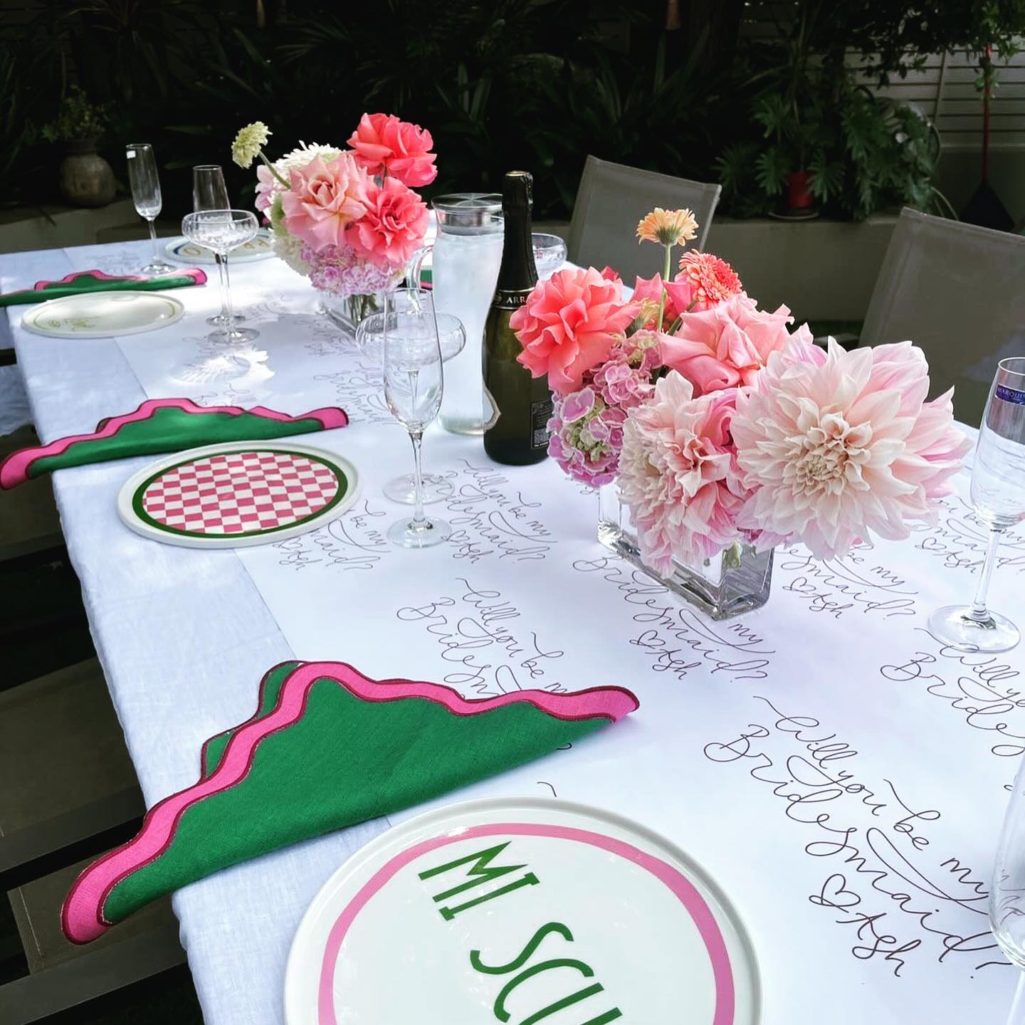 Tablescape with 'Will you be my bridesmaid'  handwritten on the table cloth decorated with bright green and pink scalloped edged napkins and pink floral centre pieces 