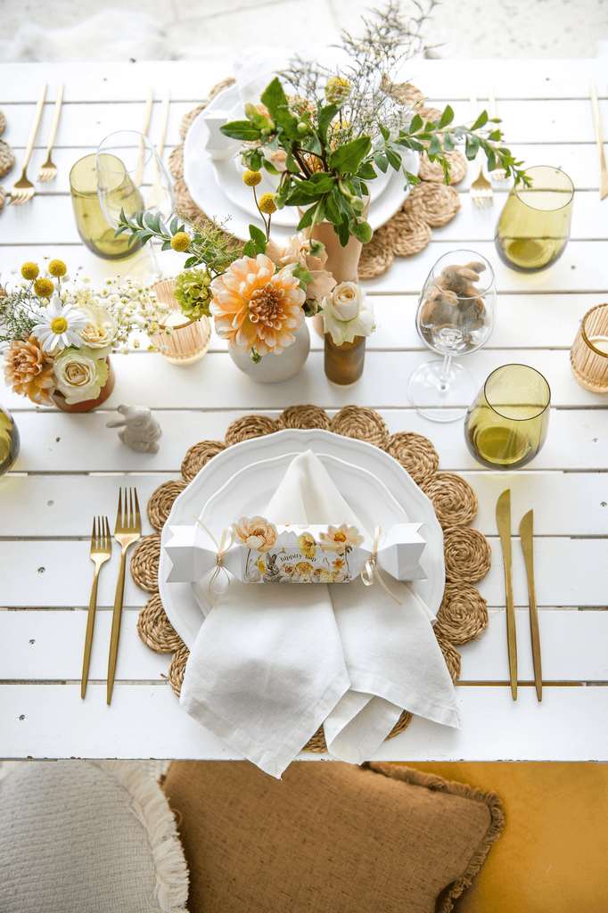 Table setting with green and yellow spring flowers, scalloped edge woven place mat, green glassware and white napkins decorated with a floral patterned cracker on top of white crockery 