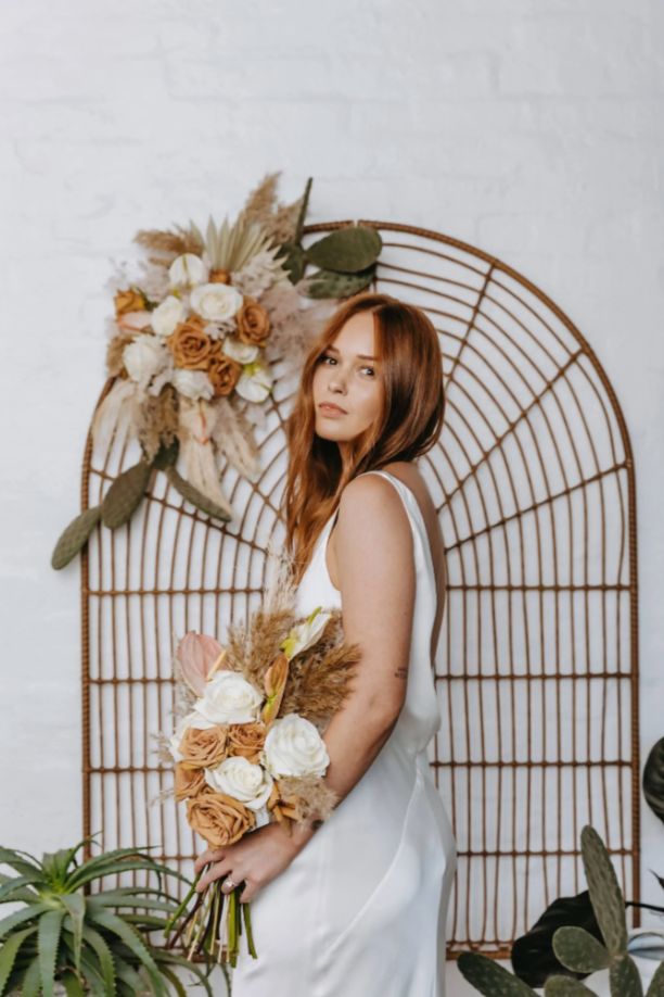 Bride holding boho bridal bouquet in front of rattan screen with boho floral arrangements