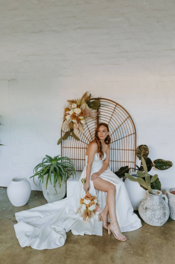 bride sits in classic white wedding gown in front of a rattan screen and greenery. Bride is holding a boho bridal bouquet