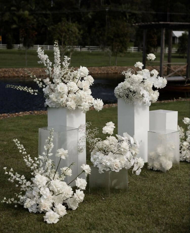 Four white square plinths stand at the top of an alter decorated with white florals for a wedding ceremony 