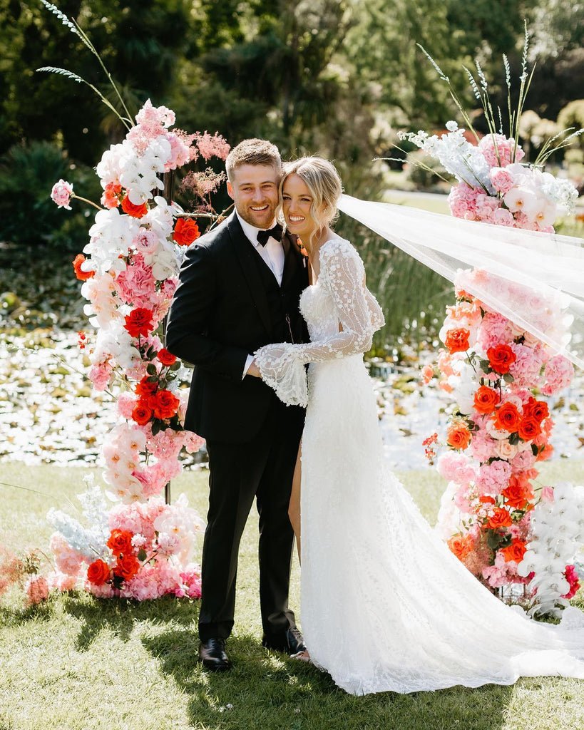 Michelle Andrews and Mitchell Rees smile as they stand side by side in front of their bright pink and red floral alter