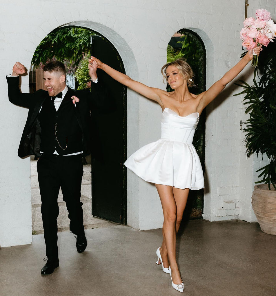 Michelle Andrews enters her wedding reception arms up in the air, wearing a satin mini party dress with husband Mitchell Rees