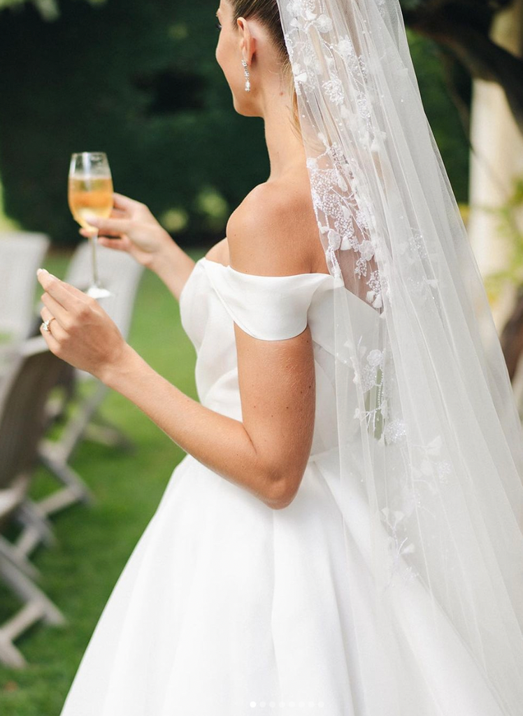 Natalie Roser Wedding. Close up photo of Natalie in white wedding dress, embroidered veil holding a glass of champagne.