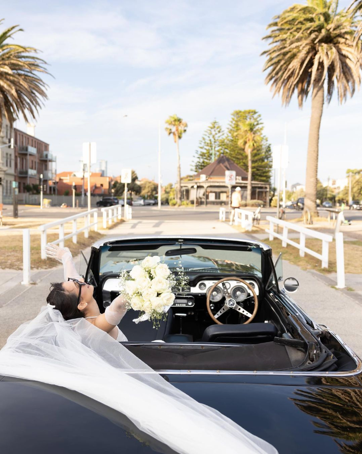 Bride sits in vintage black convertible car holding bouquet.