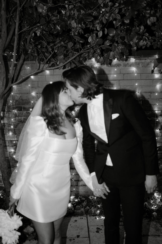 Black and white image of Bride wearing a mini wedding dress kissing a groom in a black tuxedo. 