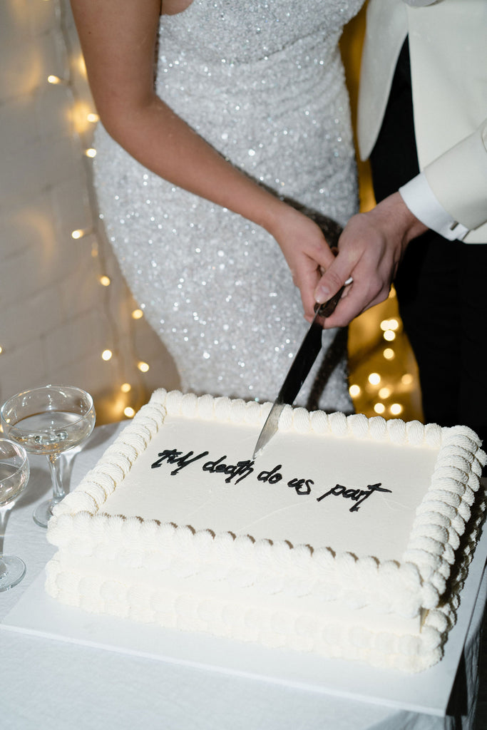 Bride and groom cut large death till us part cake standing in front of fairy lights 