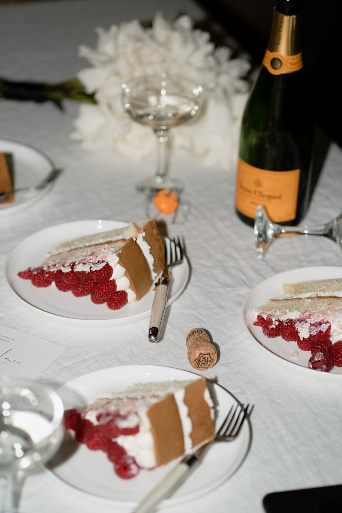 Slices of giant raspberry cake, bottle of verve champagne and a bouquet of white roses sit on a table.