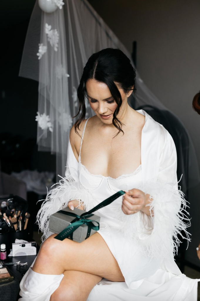 Bride sits in white robe with fluffy sleeves opening a wedding present