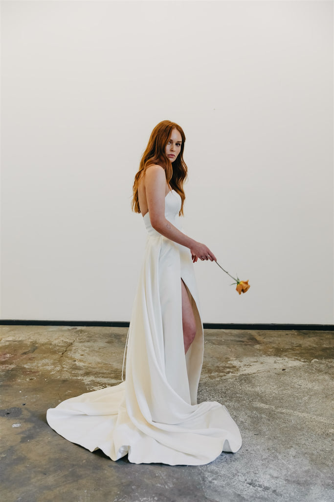 Bride stands in long wedding dress on concrete floor against white wall holding toffee rose.