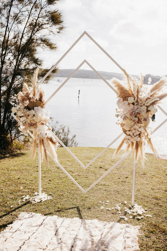 White wooden diamond arbour stands decorated with florals and a beautiful scenery of water and greenery in the background 