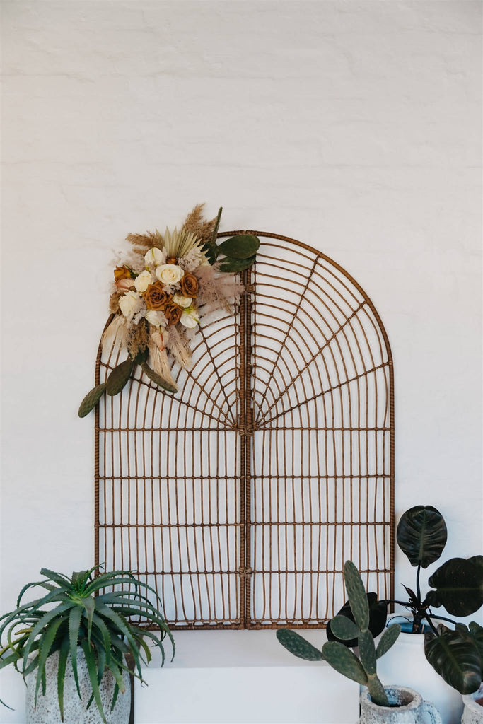 Arched rattan room divider backdrop is propped against a white wall surrounded by green plants decorating the alter of a wedding ceremony 