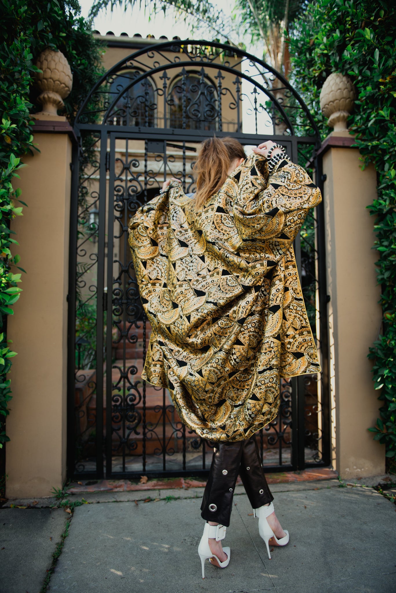 Girl in gold brocade Anthony Muto Cocoon coat leather pants & white heel front of gate