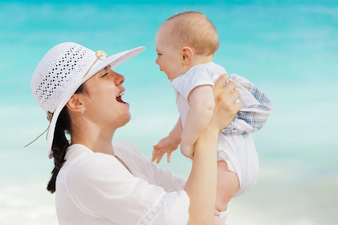 Mother wearing sun hat talking to baby she is holding up at the beach.