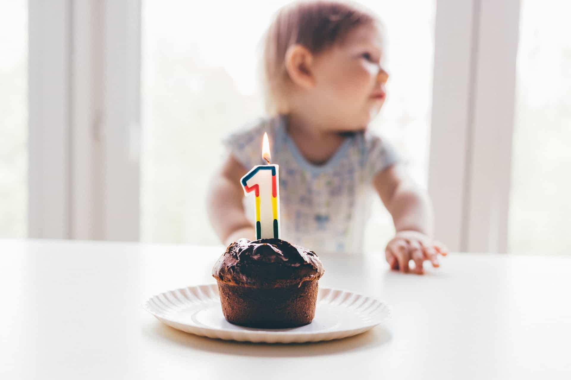 A chocolate cupcake with a “1” shaped candle and a toddler in the background.