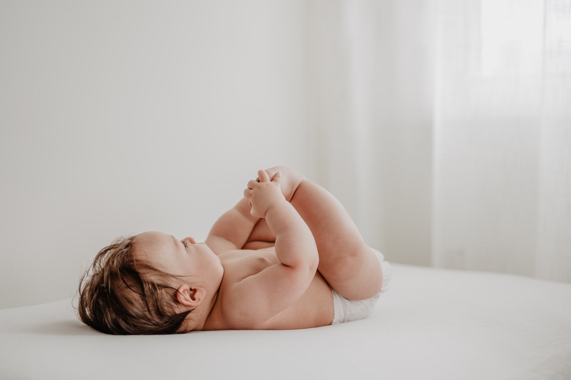Baby in diaper plays with their feet while laying on bed