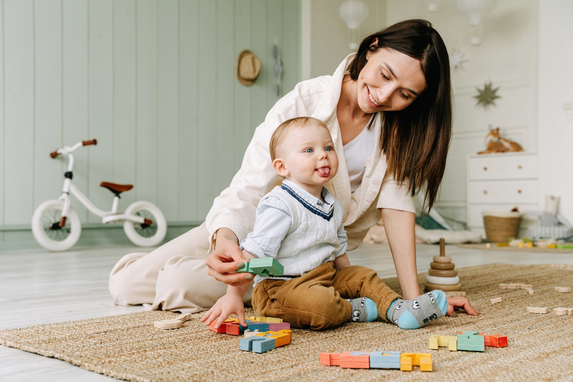 A woman sits on the floor with a baby, surrounded by toys and wooden block toys